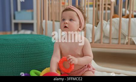 Bébé fille jouant avec un anneau de dentition rouge et des anneaux empilables colorés, tendant la main pour toucher un pouf tricoté vert à côté du berceau en bois dans la chambre d'enfant ; curiosité di Banque D'Images