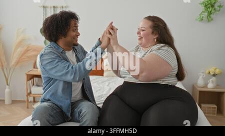 Homme et femme grande taille se donnent la main et un high five sur le lit, souriant dans une chambre confortable à l'intérieur d'un bâtiment ; connexion d'affection. Banque D'Images