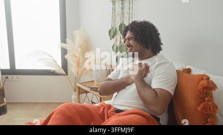 Homme serrant la poitrine sur le lit dans le bâtiment, grimacant dans la douleur sous la couverture orange portant un tshirt blanc, tenant les mains à la poitrine pour Heartattack ; urgent di Banque D'Images