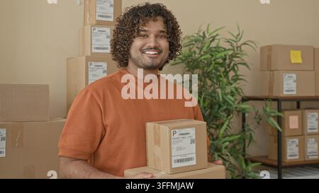 L'homme tient deux paquets de carton avec les mains, souriant et montrant l'étiquette d'expédition sur la boîte supérieure dans une zone de parcelle de bâtiment avec des boîtes empilées et une étagère Banque D'Images