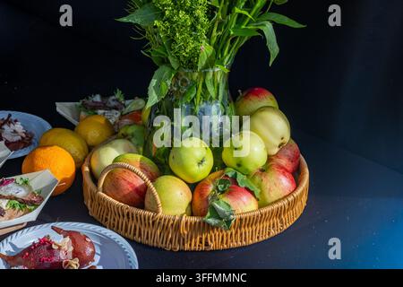 Un panier en osier rempli de pommes rouges et vertes fraîches entourant un vase en verre de feuillage vert, posé sur une table sombre à côté de plats gastronomiques et d'agrumes Banque D'Images