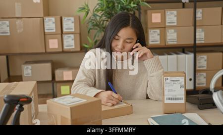 Femme écrivant une adresse sur un colis avec un stylo et sa main tout en parlant au smartphone dans un petit bâtiment commercial ; productivité. Banque D'Images