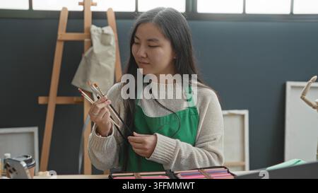 Jeune peintre tenant des pinceaux, main levée inspectant un pinceau près du chevalet et de la toile dans un studio d'art lumineux ; concentration créative. Banque D'Images