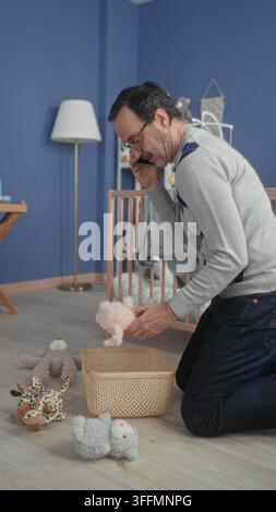 Homme au téléphone plaçant le lapin en peluche dans le panier à côté du berceau dans le bâtiment de la chambre de bébé ; organisation des soins. Banque D'Images