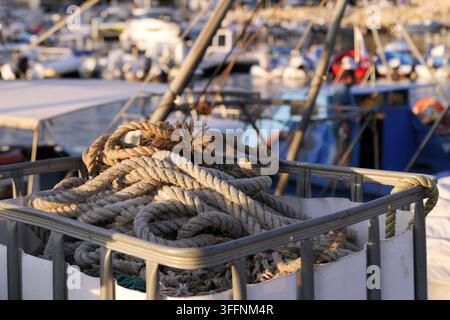 Cordes de pêche enroulées stockées dans un conteneur de bateau, fibres texturées captant la lumière du soleil avec des bateaux flous et un ouvrier portuaire en arrière-plan, maritime Banque D'Images