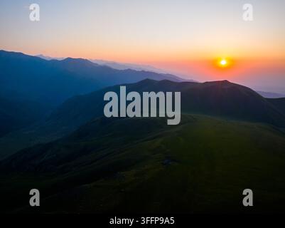 Une vue aérienne capture un paysage montagneux époustouflant au coucher du soleil au Kirghizistan. Des collines vertes ondulantes et des pics superposés créent une atmosphère sereine, rehaussée par un ciel éclatant. Banque D'Images