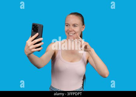 Jeune femme souriant, tenant un smartphone et faisant un geste de paix Banque D'Images