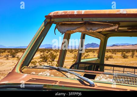 Un véhicule abandonné et rouillé est posé dans le désert sec du Mojave en Californie tandis que la lumière du soleil traverse les vitres brisées et le métal érodé. Le paysage accidenté Banque D'Images