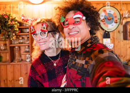 Couple souriant prend un selfie de Noël joyeux à la maison, portant des lunettes de Père Noël fantaisie et des pulls colorés dans un intérieur confortable décoré qui celebra Banque D'Images