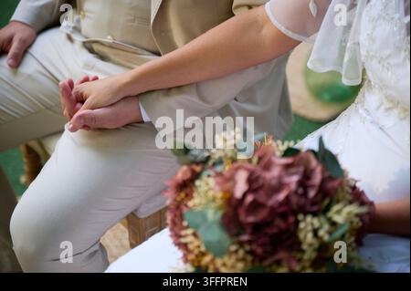 Close-up d'une mariée et d'un marié se tenant la main lors d'une cérémonie de mariage en plein air, avec un bouquet floral bordeaux reposant sur la robe blanche de la mariée. Banque D'Images