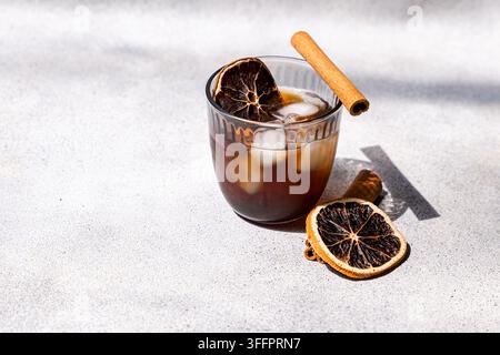 Verre de café glacé épicé avec des glaçons, un bâton de cannelle et des rondelles d'orange séchées sur une surface en pierre baignée de soleil, créant une boisson aromatique rafraîchissante Banque D'Images