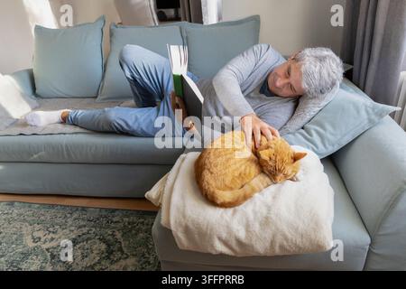 Un homme senior se détend sur un canapé confortable à la maison, lisant un livre tout en caressant doucement son chat roux reposant sur une couverture dans un salon lumineux. Banque D'Images