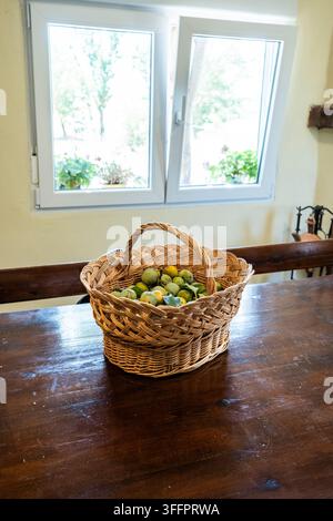 Panier tissé rempli de fruits verts frais reposant sur une table en bois sombre dans une maison rustique lumineuse, avec des vues par la fenêtre ouverte sur les arbres et la campagne au-delà Banque D'Images