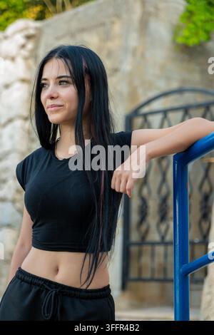 Jeune femme vêtue de noir s'appuie sur une rambarde bleue en plein air avec un léger sourire. Banque D'Images