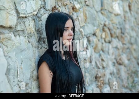 Une jeune femme aux longs cheveux noirs s'appuie contre un mur de pierre texturé en plein air. Banque D'Images