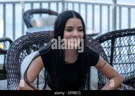 Une jeune femme aux longs cheveux foncés est assise sur une chaise en osier et sourit doucement à l'extérieur. Banque D'Images