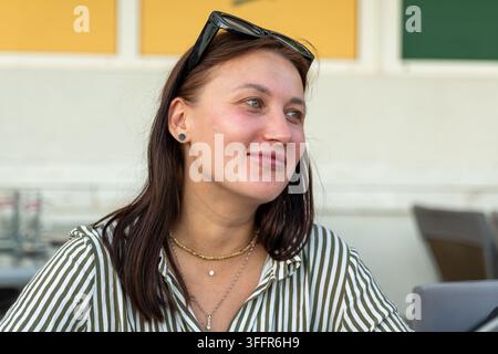 Une jeune femme souriante est assise en plein air, portant des lunettes de soleil sur la tête et une chemise à rayures. Banque D'Images