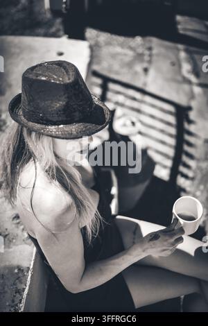 Portrait monochrome en contre-plongée d'une femme portant un chapeau fedora sombre et une robe tout en tenant une tasse, Algarve, Portugal Banque D'Images