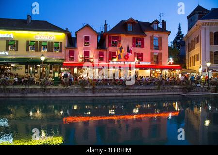 Façades de restaurants illuminées au bord de l'eau se reflétant dans les eaux calmes du canal du Le Thiou au crépuscule dans la vieille ville historique d'Annecy, France. Banque D'Images
