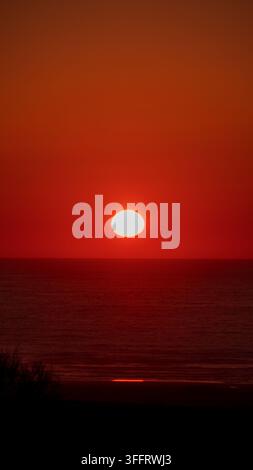 Prise verticale du disque solaire complet se couchant dans un ciel rouge intense au crépuscule au-dessus de l'horizon calme de l'océan Atlantique au Maroc Banque D'Images