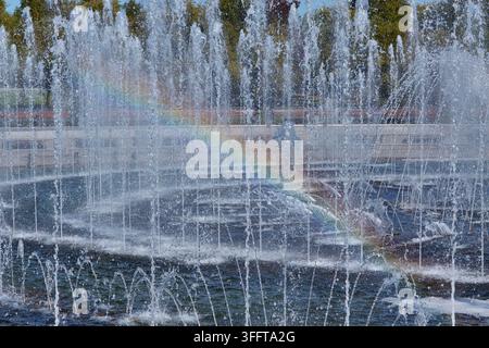 Fontaine à eau affichant plusieurs jets et un léger arc-en-ciel dans un parc, avec des arbres et un ciel lumineux visibles en arrière-plan Banque D'Images