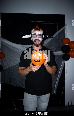 Homme souriant en maquillage d'Halloween et costume d'horreur tenant un ballon orange amusant décoré d'un visage de monstre. Banque D'Images