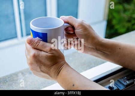 Gros plan de mains âgées tenant une tasse de café blanche près d'une fenêtre ouverte, montrant une pause matinale tranquille à la maison. Banque D'Images