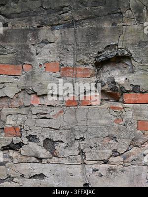 Surface de mur enduite de ciment endommagée avec brique rouge apparente, enduit à arêtes fissuré, sangles métalliques, grande ouverture murale et couche sous-jacente sombre Banque D'Images