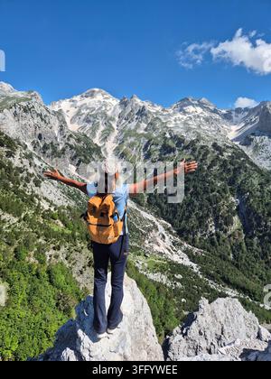Un voyageur se tient triomphalement sur une corniche rocheuse à Teth, Albanie, entouré de vues montagneuses à couper le souffle. La lumière du soleil baigne le paysage accidenté, hig Banque D'Images