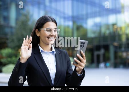 Femme d'affaires debout à l'extérieur devant un bâtiment de bureaux moderne, souriant et agitant la main tout en passant un appel vidéo sur un smartphone, se connectant et communiquant à distance Banque D'Images