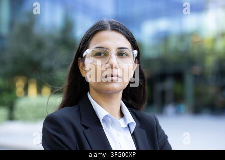 Jeune femme d'affaires indienne portant un costume et des lunettes transparentes, debout avec assurance en plein air devant un bâtiment moderne en verre, incarnant le professionnalisme et le leadership d'entreprise Banque D'Images