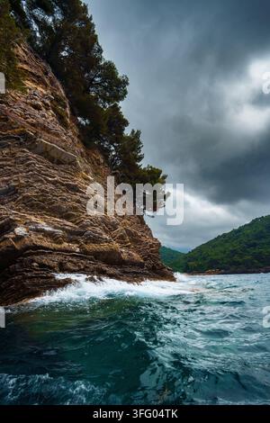 Prise verticale d'une falaise escarpée et stratifiée rencontrant les vagues turquoise de la mer sous un ciel orageux à Petrovac, au Monténégro. Banque D'Images