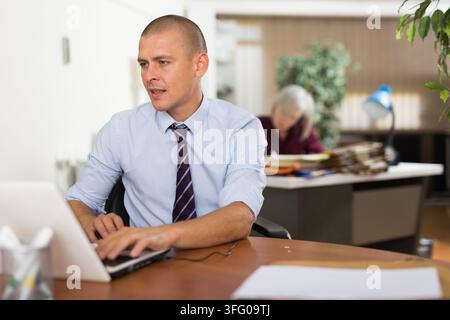 Un employé de bureau assis à une table et utilisant son ordinateur portable. Une femme aux cheveux gris assise en arrière-plan.. Banque D'Images