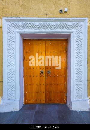 L’entrée majestueuse présente de hautes doubles portes en bois ornées de clous en laiton et de heurtoirs en tête de lion. Encadrée par un portail en pierre sculptée contre un mur jaune texturé. Banque D'Images