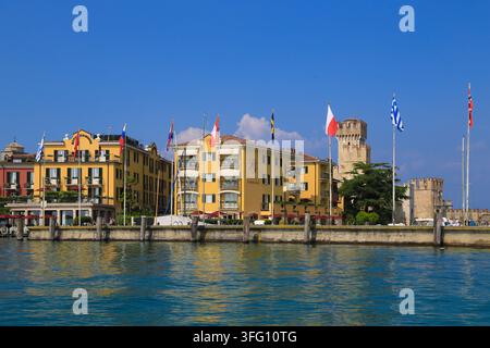 Hôtels jaunes vifs au bord du lac arborant des drapeaux internationaux à côté des tours en pierre historiques du château Scaligero sur le lac de Garde à Sirmione, en Italie. Banque D'Images