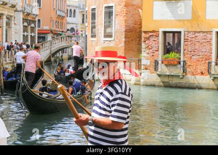 Un gondolier vénitien joyeux portant une chemise à rayures et un chapeau de paille pose sur un canal calme alors que des gondoles transportent des touristes sous un pont de pierre à Venise. Banque D'Images