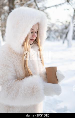 Jeune femme tient une tasse réutilisable chaude avec du café dans un parc enneigé, profitant d'une pause calme et confortable pendant une journée d'hiver froide. Format vertical. Banque D'Images