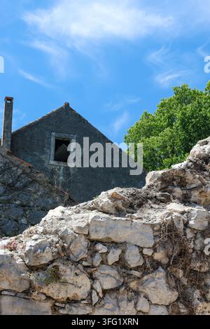 Une maison en pierre abandonnée et un mur de roche en ruine se dressent sous un ciel bleu vif et des nuages filamenteux à côté d'un arbre vert feuillu, évoquant une décadence rustique, soli Banque D'Images