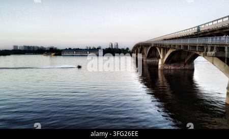 Bateau à moteur filant sur une large surface fluviale près d'une structure massive de pont en arche en béton. Vue grand angle représentant le transport fluvial actif et urbain c Banque D'Images