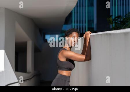 Une femme noire repose ses bras sur un mur après l'exercice. Une femme afro-américaine semble concentrée et composée dans un environnement de fitness moderne en plein air. Banque D'Images
