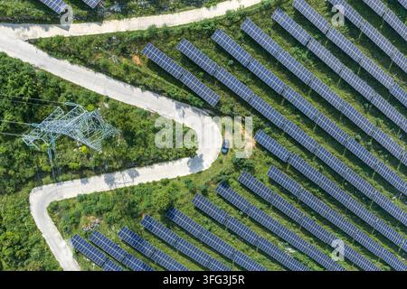 Une prise aérienne montre des rangées de panneaux solaires bien ordonnées sur des collines herbeuses, traversées par une route sinueuse et une tour d'alimentation en acier sous une lumière du jour éclatante. Banque D'Images