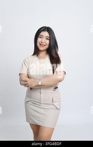 Portrait d'une jeune femme asiatique souriante en tenue beige décontractée, debout les bras croisés sur un fond blanc de studio Banque D'Images