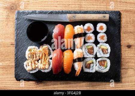 Assortiment de sushi japonais servi sur une planche d'ardoise noire avec des nigiri au saumon et au thon, des crevettes, des rouleaux maki, de la sauce soja et un couteau de cuisine sur une table en bois. Banque D'Images
