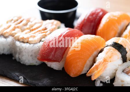 Assortiment de sushi japonais servi sur une planche d'ardoise noire avec des nigiri au saumon et au thon, des crevettes, des rouleaux maki, de la sauce soja et un couteau de cuisine sur une table en bois. Banque D'Images