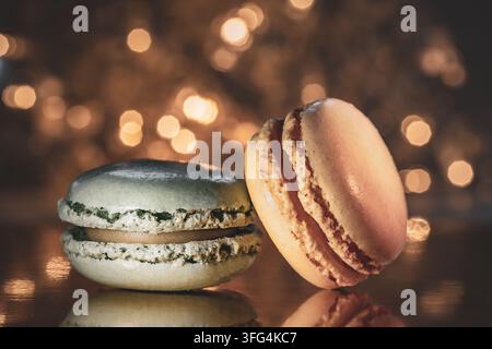 Deux délicieux macarons français reposant sur une surface réfléchissante devant un fond bokeh chaleureux et festif de guirlandes lumineuses. Banque D'Images