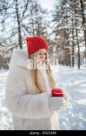 Jeune femme tient une boisson chaude à emporter avec les deux mains dans un parc enneigé, se réchauffant lors d'une promenade hivernale froide et profitant d'une pause saisonnière confortable. Verti Banque D'Images