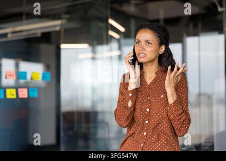 Jeune femme d'affaires communiquant sur un téléphone mobile avec une expression contrariée, gesticulant de la main dans un bureau moderne, subissant du stress et des préoccupations professionnelles Banque D'Images