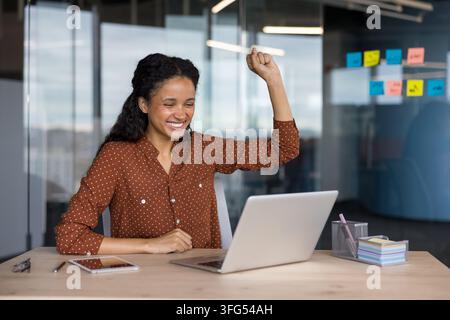 Femme d'affaires enthousiaste applaudissant avec un bras levé tout en regardant l'écran de son ordinateur portable, ressentant un sentiment d'accomplissement et de réussite à son espace de travail moderne Banque D'Images