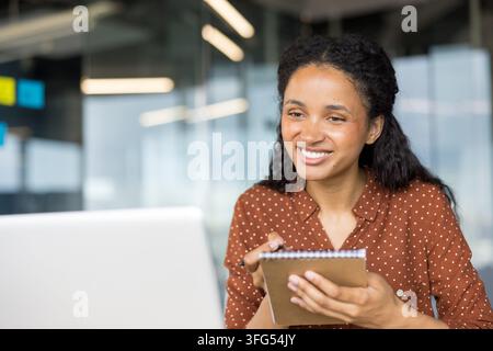 Femme souriante, prenant des notes dans un cahier à spirale tout en travaillant à distance sur un ordinateur portable, participant activement à une réunion virtuelle ou à une session d'apprentissage en ligne Banque D'Images