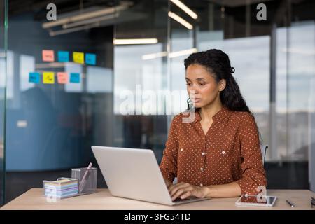 Jeune femme professionnelle se concentrant sur son ordinateur portable, tapant à un bureau en bois dans un environnement de bureau en verre contemporain, entourée de notes transparentes colorées sur le mur Banque D'Images